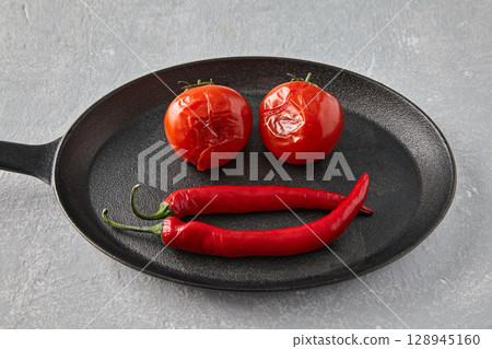 baked tomatoes and hot peppers in a black cast iron skillet on a grey stone table. 128945160