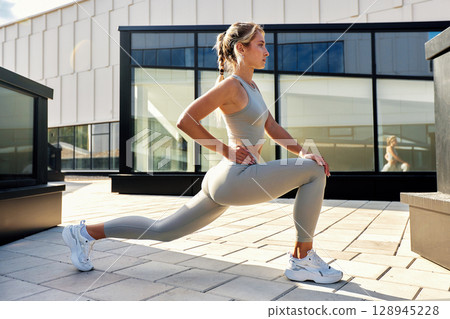 Slender blonde sportswoman doing exercises against the blue summer sky and gray modern building background. Young female athlete enjoys morning workouts. Regular trainings in urban environment. Slender blonde sportswoman doing exercises against the blue summer sky and gray modern building background. Young female athlete enjoys morning workouts. Regular trainings in urban environment. 128945228