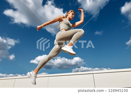 Young female athlete doing outside workout on a sunny day. Slender blonde girl in gray sportswear makes jumps, does functional training in the street. Active lifestyle in urban environment. Young female athlete doing outside workout on a sunny day. Slender blonde girl in gray sportswear makes jumps, does functional training in the street. Active lifestyle in urban environment. 128945230