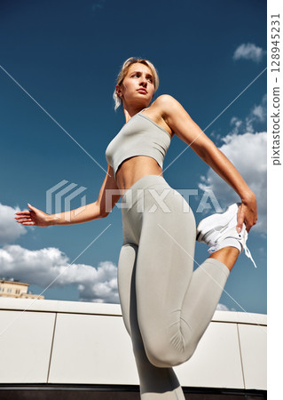 Young female athlete doing outside workout on a sunny day. Slender blonde girl in gray sportswear makes jumps, does functional training in the street. Active lifestyle in urban environment. Young female athlete doing outside workout on a sunny day. Slender blonde girl in gray sportswear makes jumps, does functional training in the street. Active lifestyle in urban environment. 128945231