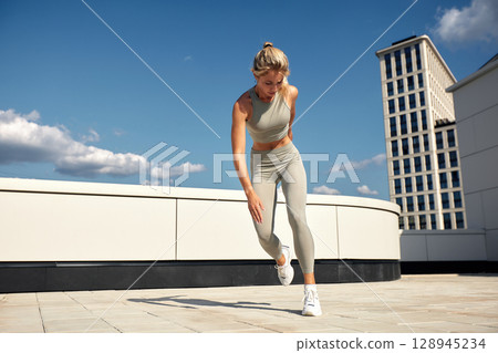 Young female athlete doing outside workout on a sunny day. Slender blonde girl in gray sportswear makes jumps, does functional training in the street. Active lifestyle in urban environment. Young female athlete doing outside workout on a sunny day. Slender blonde girl in gray sportswear makes jumps, does functional training in the street. Active lifestyle in urban environment. 128945234
