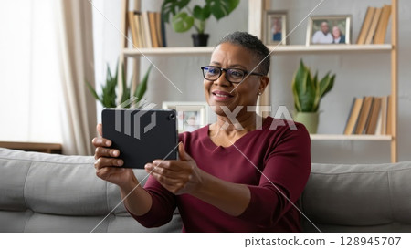 Cheerful senior woman video chatting on digital tablet in cozy home setting with bookshelves. Generative AI. 128945707