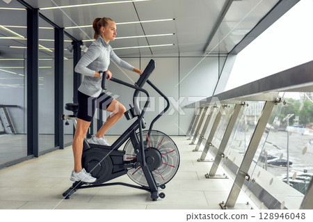 Woman exercising on a modern air bike in a sleek gym interior Woman exercising on a modern air bike in a sleek gym interior 128946048
