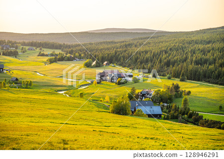 A serene evening unfolds in Jizerka Village, nestled in the Jizera Mountains. The golden sunlight casts a warm glow over lush green fields and charming houses, creating a peaceful atmosphere. 128946291
