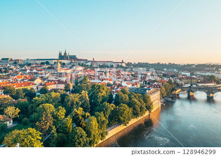 Prague awakens with the golden hues of sunrise illuminating the medieval architecture. The Vltava River reflects the breathtaking view of the city, including the iconic Charles Bridge and castle. 128946299