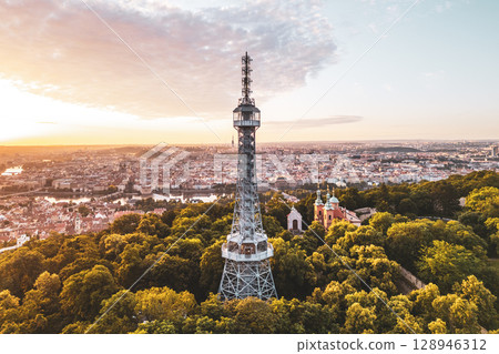 Pink and orange hues fill the sky as the sun rises over the Petrin Lookout Tower in Prague. The tower stands tall among lush greenery, offering a breathtaking view of the city skyline. Pink and orange hues fill the sky as the sun rises over the Petrin Lookout Tower in Prague. The tower stands tall among lush greenery, offering a breathtaking view of the city skyline. 128946312