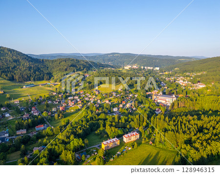 Scenic view of Tanvald nestled in the Jizera Mountains, showcasing lush greenery, charming houses, and rolling hills under a clear blue sky. A perfect summer day highlights this serene landscape. Scenic view of Tanvald nestled in the Jizera Mountains, showcasing lush greenery, charming houses, and rolling hills under a clear blue sky. A perfect summer day highlights this serene landscape. 128946315