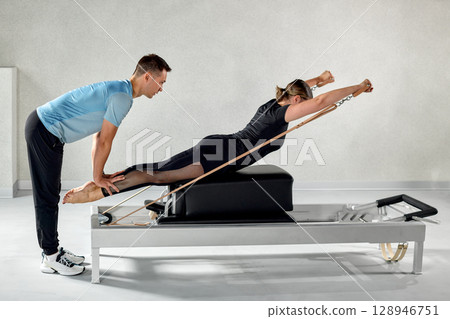 Woman practicing Pilates with assistance on equipment in fitness studio 128946751