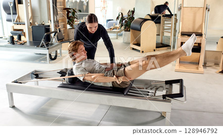 Trainer assisting individual with pilates exercise on reformer equipment in bright studio 128946792