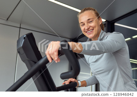 Smiling woman exercising on elliptical trainer indoors at modern fitness center Smiling woman exercising on elliptical trainer indoors at modern fitness center 128946842