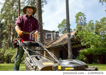 Man mowing lawn with a gas-powered mower wearing gloves and sunhat Man mowing lawn with a gas-powered mower wearing gloves and sunhat 128946856
