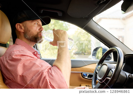 Man drinking water inside a car with a focused expression and casual outfit 128946861