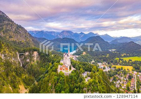 Perched on a hilltop, Neuschwanstein Castle emerges from lush greenery and mountains, revealing its fairytale beauty against a backdrop of serene lakes and dramatic skies. 128947114
