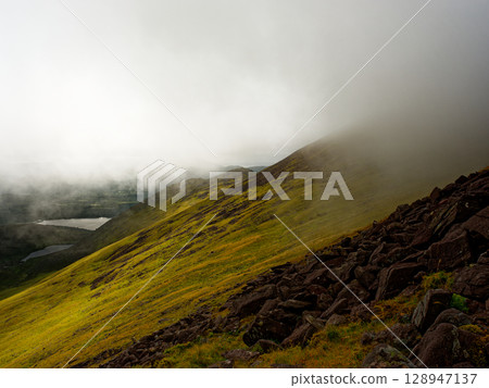 Green hillsides meet rocky terrain in the Coomloughra Horseshoe, Ireland. Low clouds and mist obscure the mountain peaks, creating a moody atmosphere with visible lakes below. 128947137