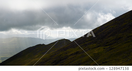 The slopes of Coomloughra Horseshoe mountains rise towards a cloudy sky. Sunlight filters through the clouds, illuminating the landscape in County Kerry, Ireland. 128947138