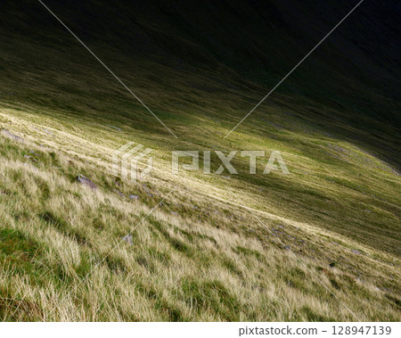 A sunlit grassy hillside in the Coomloughra Horseshoe, Ireland, shows contrasting patches of light and shadow. The steep slope leads upwards to a dark ridge. 128947139