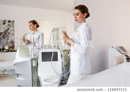 Professional cosmetologist in white uniform with modern aesthetic laser machine for non-invasive skin treatment in beauty clinic or medical spa, advanced cosmetic equipment in clean bright interior Professional cosmetologist in white uniform with modern aesthetic laser machine for non-invasive skin treatment in beauty clinic or medical spa, advanced cosmetic equipment in clean bright interior 128947315