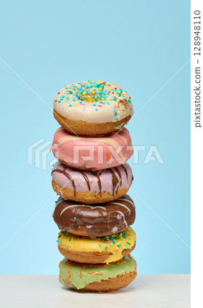 A tall stack of six colorful, glazed donuts with various toppings is centrally placed on a white surface against a solid light blue background. A tall stack of six colorful, glazed donuts with various toppings is centrally placed on a white surface against a solid light blue background. 128948110
