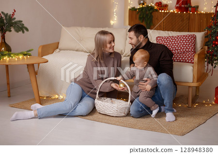 Happy young family with toddler playing on floor near sofa in cozy room with Christmas lights Happy young family with toddler playing on floor near sofa in cozy room with Christmas lights 128948380