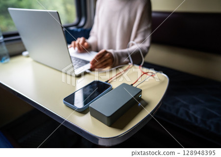 Woman working on laptop, having it and phone connected to power bank, sitting in train compartment. Woman working on laptop, having it and phone connected to power bank, sitting in train compartment. 128948395