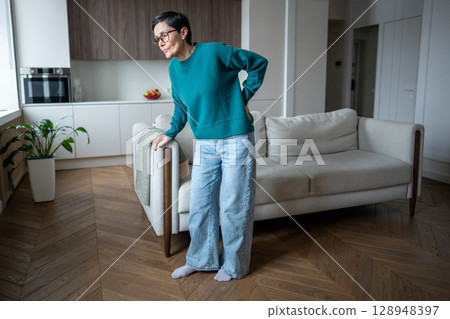 Bent over mature woman with severe lumbar pain holding couch. Back inflammation, nerve compression 128948397