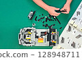 A child's hands assembling a LEGO car model. Various colorful pieces are scattered on a green surface.  128948711