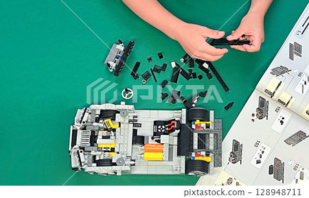 A child's hands assembling a LEGO car model. Various colorful pieces are scattered on a green surface. A child's hands assembling a LEGO car model. Various colorful pieces are scattered on a green surface. 128948711