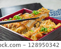 A lacquered Japanese bento box features a crisp pork cutlet glazed in savory sauce and garnished with spring onions, freshly picked up by chopsticks to highlight the dish in a menu photo. 128950342