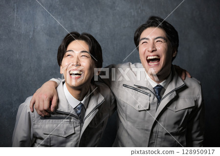 灰色背景中，兩位穿著工作服、笑容滿面的男士互相摟著肩膀站著 128950917