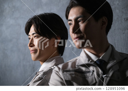 Close-up of two men standing back to back in work clothes with serious expressions on a gray background 128951096