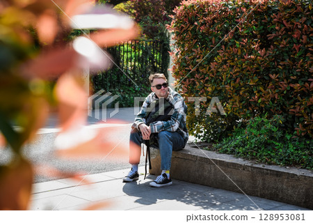 Russian male tourist resting on a stone bench near autumn shrubs in a city park, enjoying a sunny day during his travels. 128953081