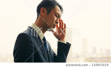 A male business person worries while looking at the city from the office window A male business person worries while looking at the city from the office window 128953819