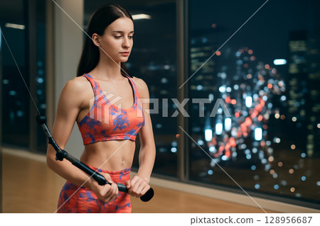 AI generated image of a fit woman in a vibrant sports outfit performing a tricep workout in a modern gym with a cityscape at night. Clean, bright, and focused on health and strength. 128956687