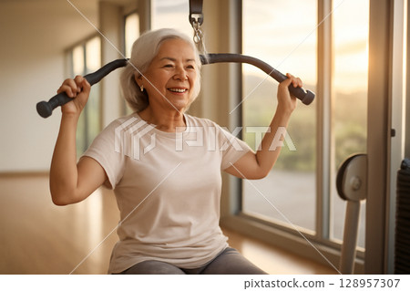 AI generated image of an elderly Asian woman in a gym working on a lat pulldown machine. She has gray hair and a warm smile, showing strength and vitality. The scene captures a peaceful, clean gym 128957307