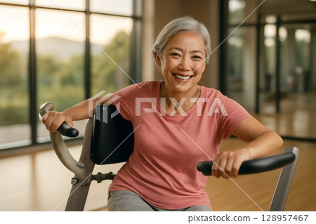 AI generated image of an elderly Asian woman in her 60s exercising in a spacious gym with natural light. She is smiling while using equipment, promoting healthy aging and well-being. AI generated image of an elderly Asian woman in her 60s exercising in a spacious gym with natural light. She is smiling while using equipment, promoting healthy aging and well-being. 128957467
