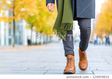 In an urban autumn setting, a stylish man walks confidently, dressed in a dark gray overcoat, brown boots, and an olive scarf, surrounded by golden leaves and modern architecture. 128959632