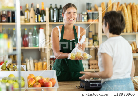 Young woman seller weighs apples in bag 128959960