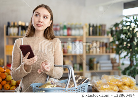 Young woman checking phone during shopping in grocery store 128960494