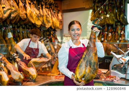 Cheerful girl owner of butcher shop holding leg of dry-cured Iberian ham 128960846