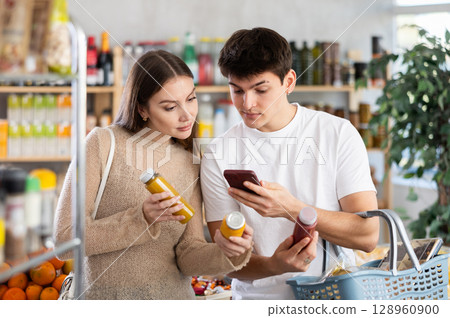 Young happy couple buyers carefully examines and view bottle of smoothie, scan QR in supermarket 128960900