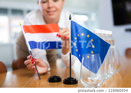 Young woman putting flags of Netherlands and EU on table in office Young woman putting flags of Netherlands and EU on table in office 128960934
