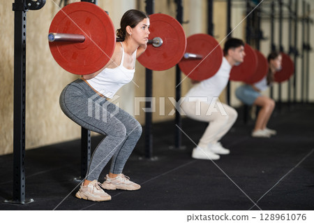 Girl in activewear exercising with barbell during group workout in gym center 128961076