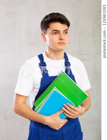 Positive worker in blue jumpsuit poses with large stack of notebooks. Positive worker in blue jumpsuit poses with large stack of notebooks. 128961083