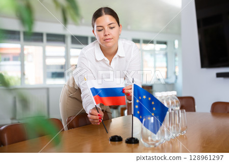 Woman secretary prepares an office for negotiations - she places flags of European Union and Russia table Woman secretary prepares an office for negotiations - she places flags of European Union and Russia table 128961297
