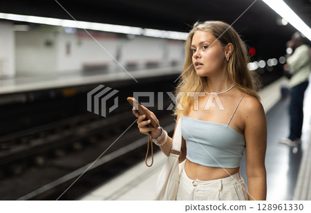 Girl with phone is standing on subway platform, waiting for train to arrive Girl with phone is standing on subway platform, waiting for train to arrive 128961330