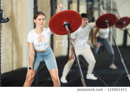 Athletic young woman pumping up her arm biceps using one side of barbell Athletic young woman pumping up her arm biceps using one side of barbell 128961451