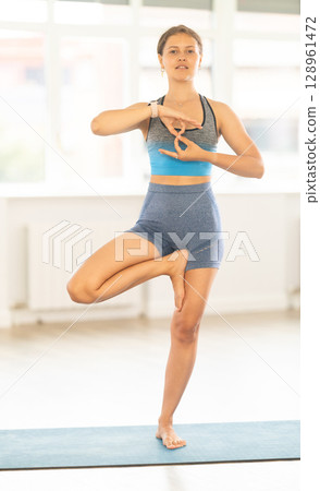 Young girl in sportswear exercising at yoga studio, standing in warrior one leg pose on fitness mat Young girl in sportswear exercising at yoga studio, standing in warrior one leg pose on fitness mat 128961472