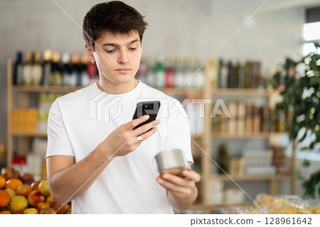 Young man customer at shop scans QR code on tin cans of preserves using phone camera Young man customer at shop scans QR code on tin cans of preserves using phone camera 128961642