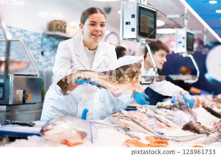 Female seller of fish department of a supermarket demonstrates fresh atlantic cod Female seller of fish department of a supermarket demonstrates fresh atlantic cod 128961733