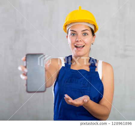 Smiling female worker in work clothes and hardhat demonstrating screen of her phone 128961896